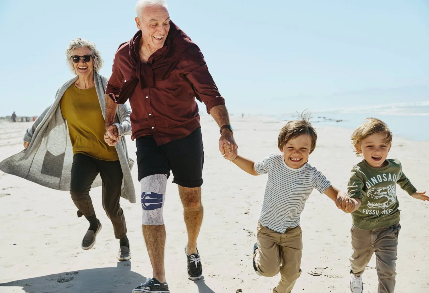 Grandparents running with their grandchildren on the beach while holding hands. The grandfather is wearing a Bauefeind GenuTrain Knee Brace to help relieve early warning symptoms of knee arthritis