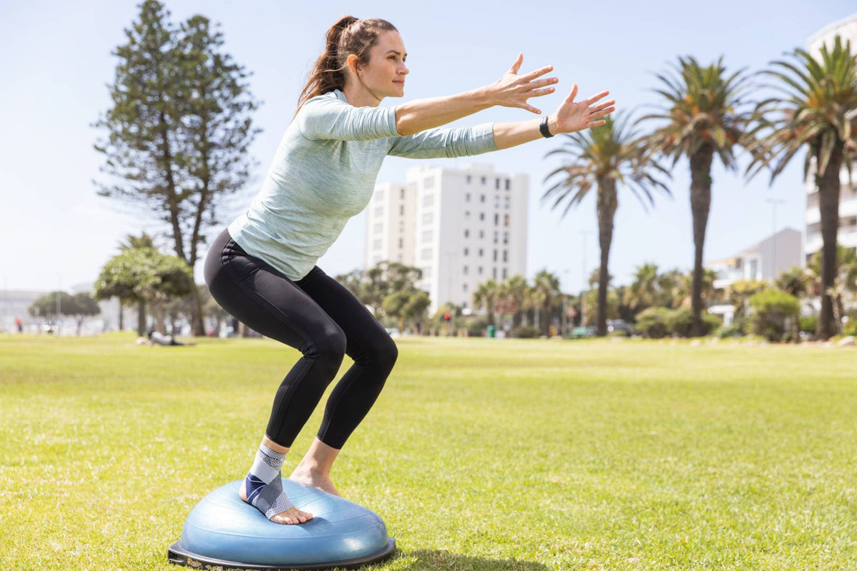 Woman in the park doing squats on a balance ball while wearing a Bauerfeind ankle brace to help with her ankle tendonitis