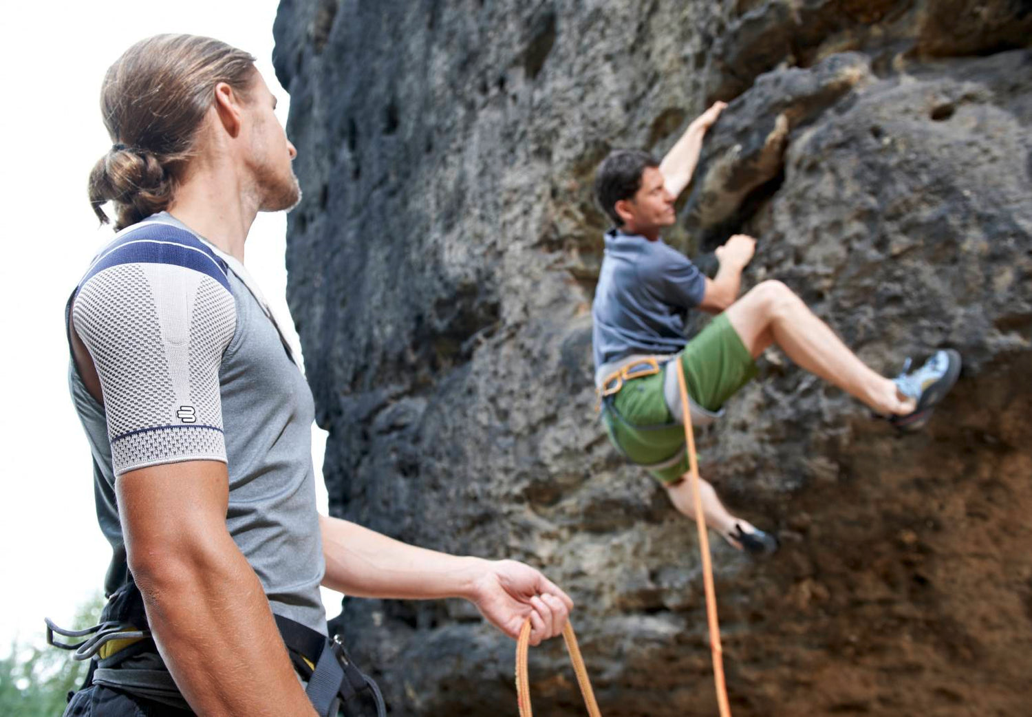 Man holding the rope for his friend while he mountain climbs. The man is wearing an OmoTrain S Shoulder Brace to manage shoulder pain