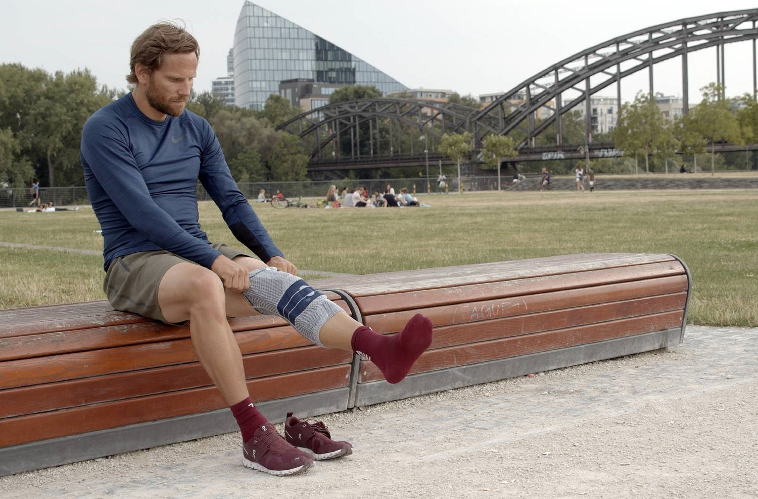 Man sitting on a bench at the park in winter pulling on Bauerfeind's GenuTrain knee brace