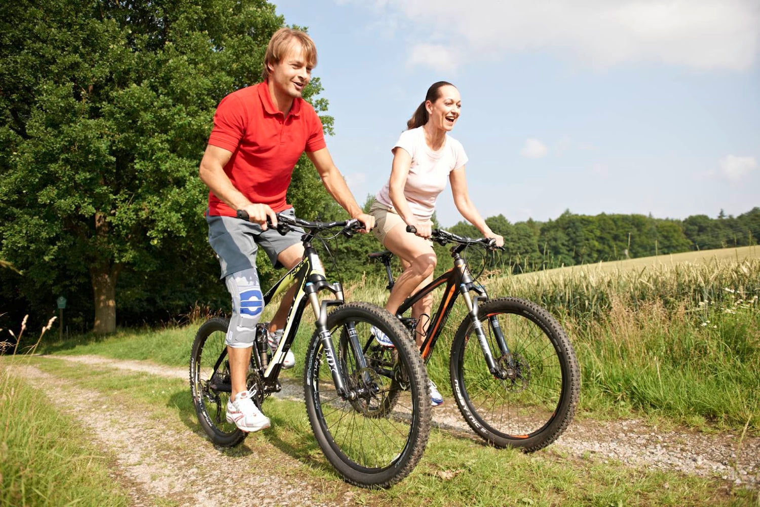 Man and woman cycling through a grassy field on a sunny day. The man is wearing Bauerfeind's SofTec Genu Kne Brace to assist in his return to cycling after injury