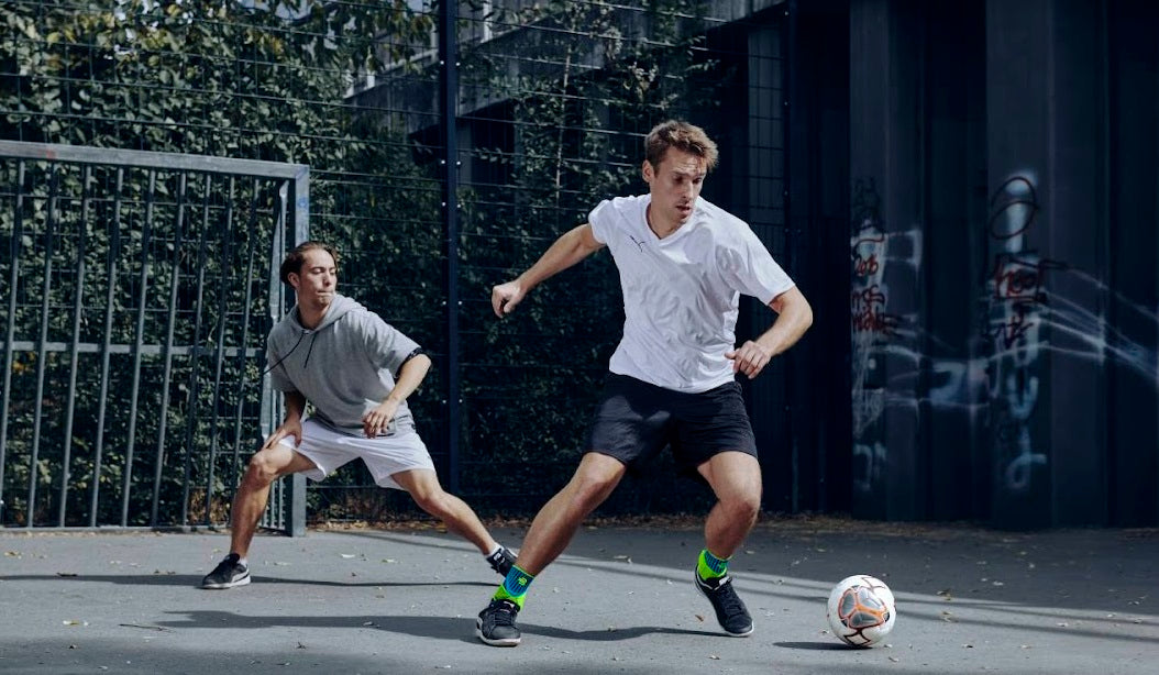 two men playing soccer on a concrete court. The one in front is Bauerfeind's sports ankle compression sleeves