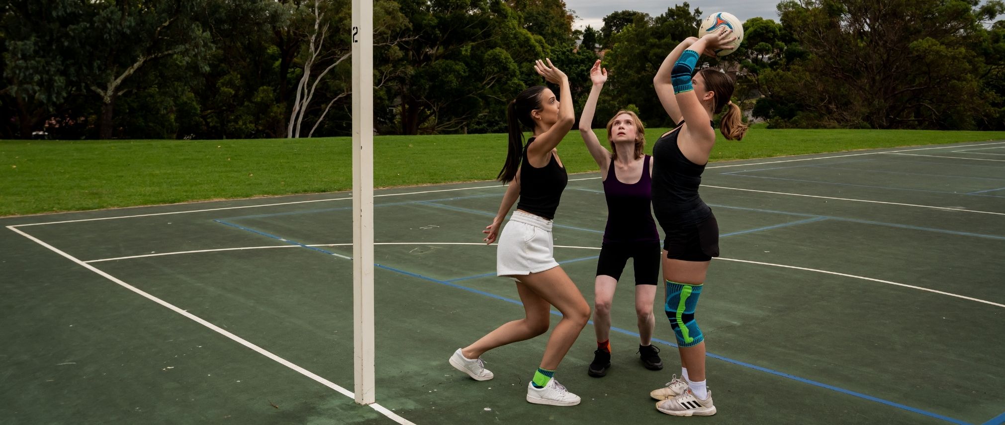 3 girls playing netball. One is trying to throw the ball into the hoop while the others are trying to block her. The girl is wearing Bauerfeind's Sports Knee Support.