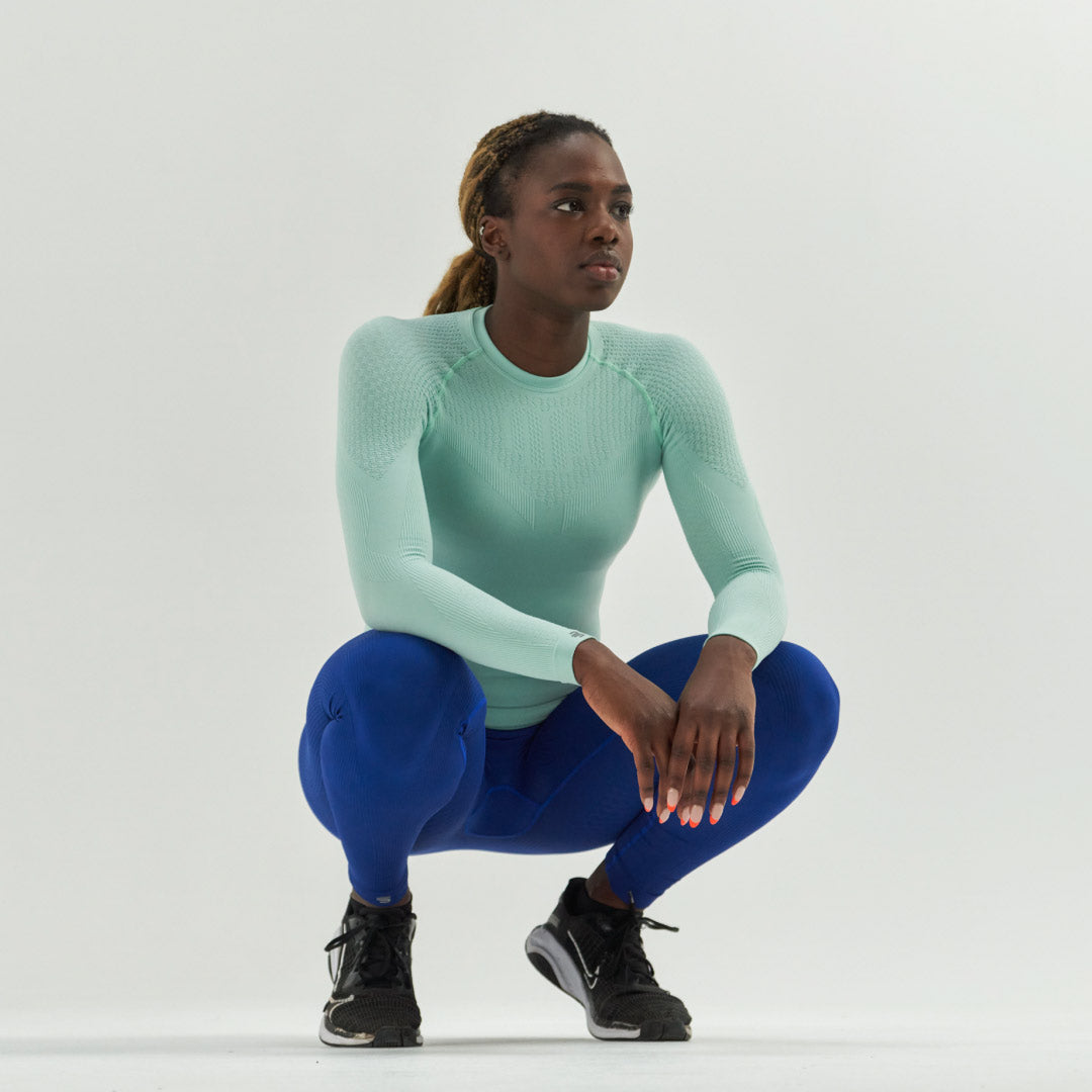 Woman in teal Bauerfeind compression long-sleeve top and blue leggings squatting on a white background