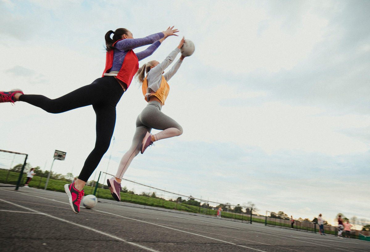 Two women jumping up to catch a netball. One is wearing a red bib and the other a yellow bib