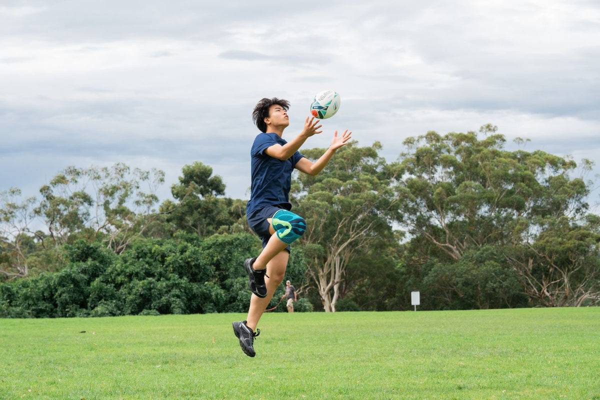 young man playing rugby in the park jumping up to catch the ball while wearing Bauerfeind's Sports Knee Support