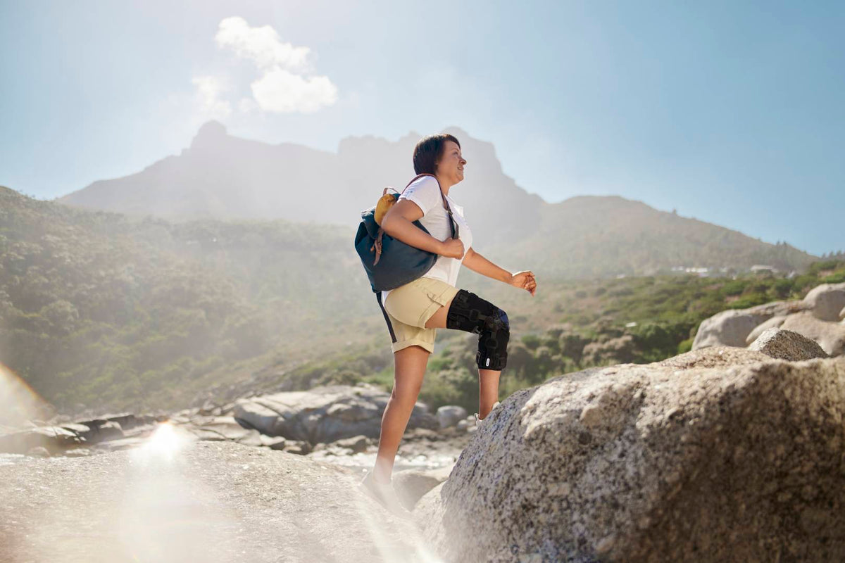 Woman walking along rocks at the beach on a sunny day. She is carrying a backpack and wearing Bauerfeind's GenuTrain OA Osteoarthritis knee brace