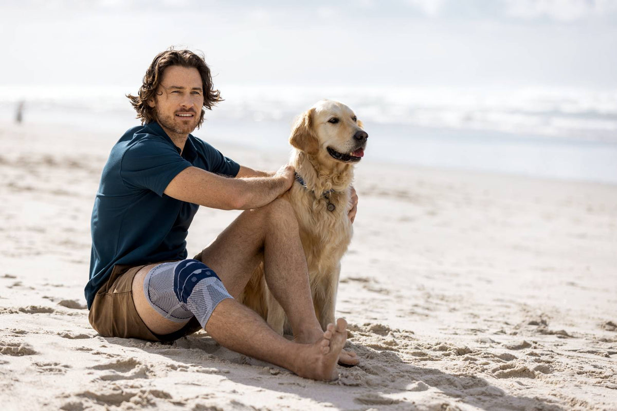 Man sitting with his dog at the beach. He is wearing the GenuTrain knee brace to help with his knee clicking pain