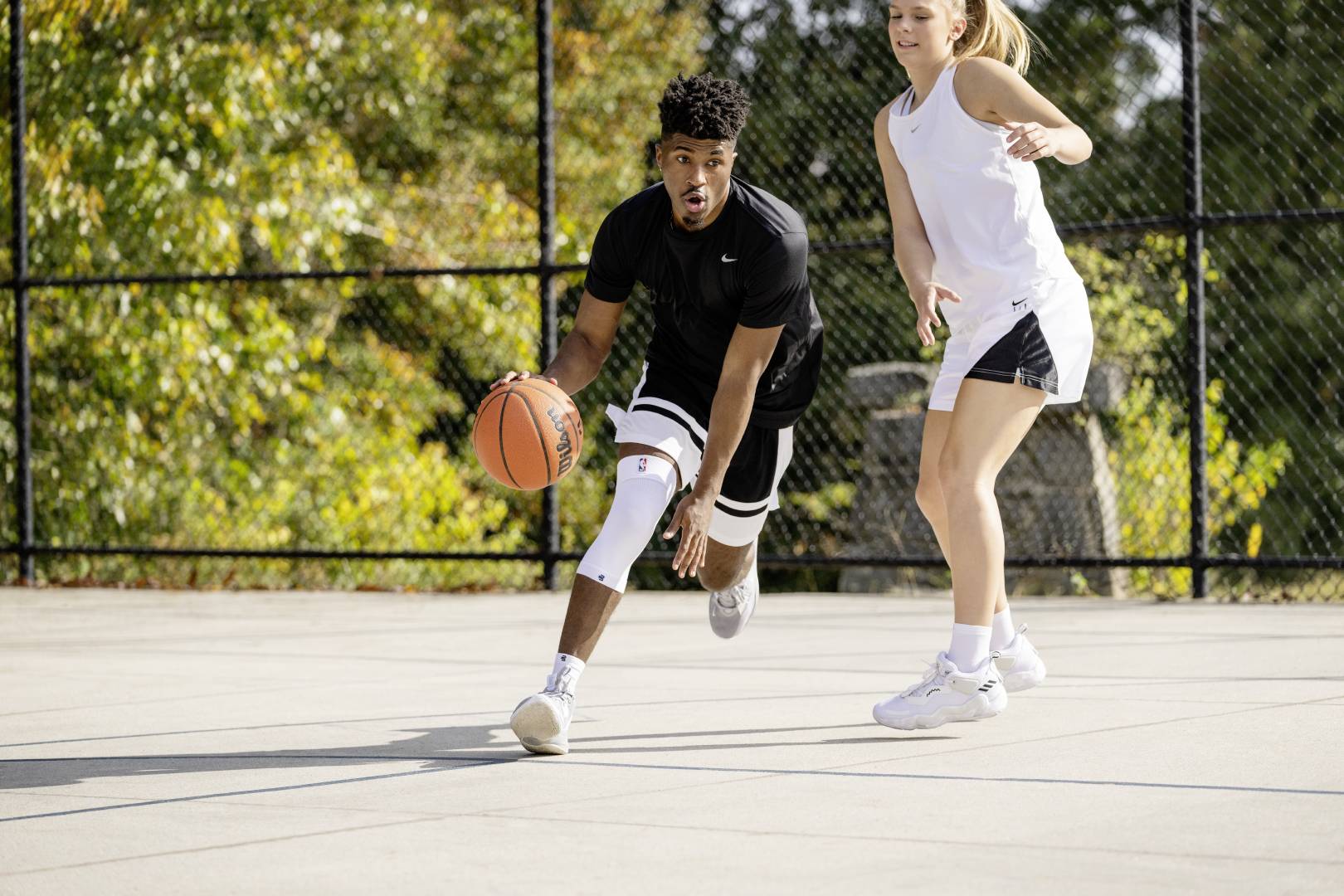 2 people playing basketball on an outdoor court. The man is wearing Bauerfeind NBA Sports compression knee support to protect his knee from injury