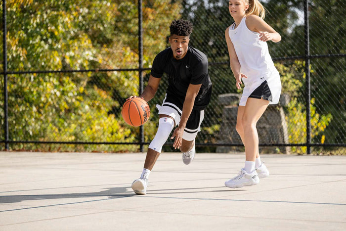 2 people playing basketball on an outdoor court. The man is wearing Bauerfeind NBA Sports compression knee support to protect his knee from injury
