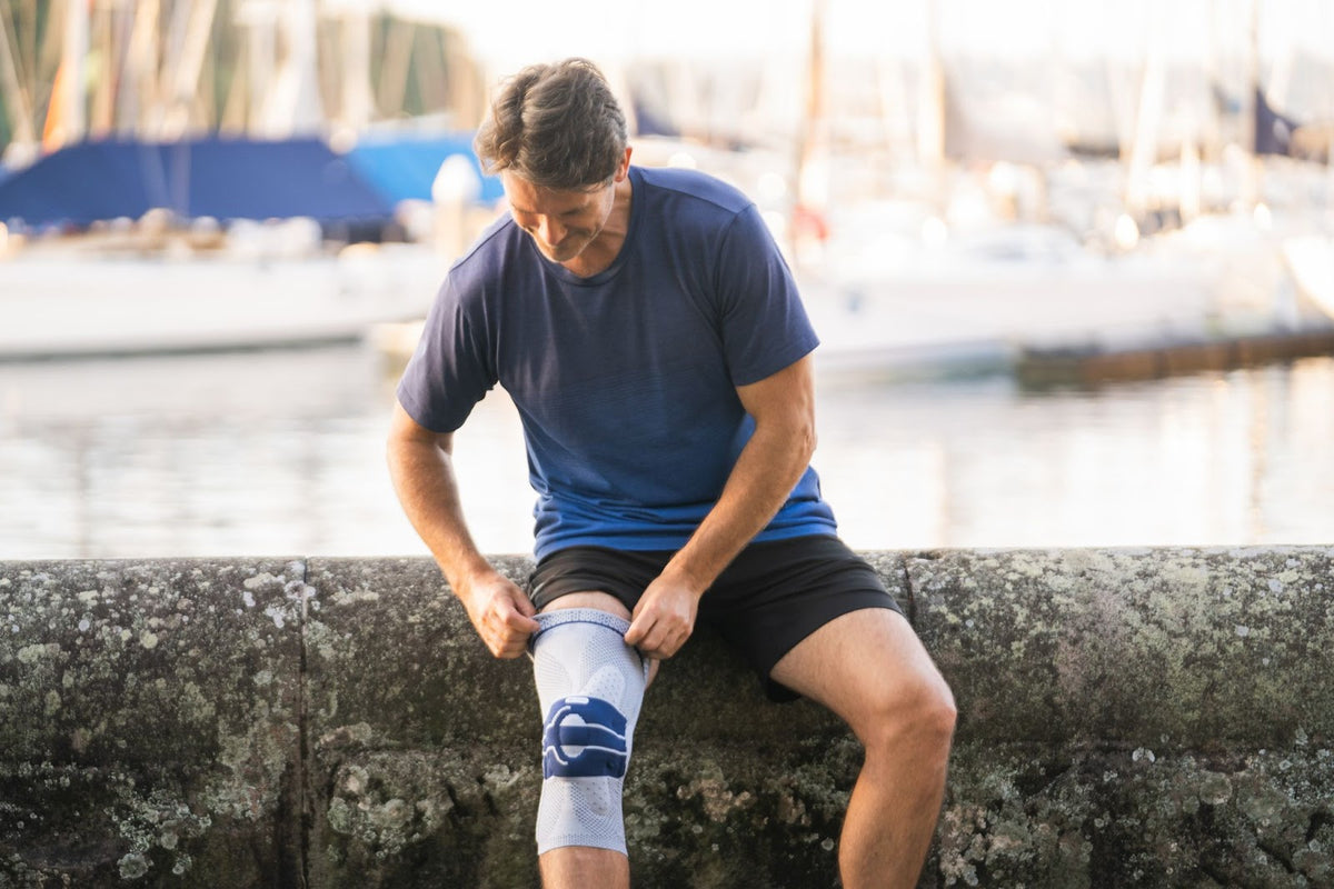 Man sitting on a stone railing at the wharf. He is pulling on a Bauerfeind GenuTrain Knee Brace to help manage knee pain 