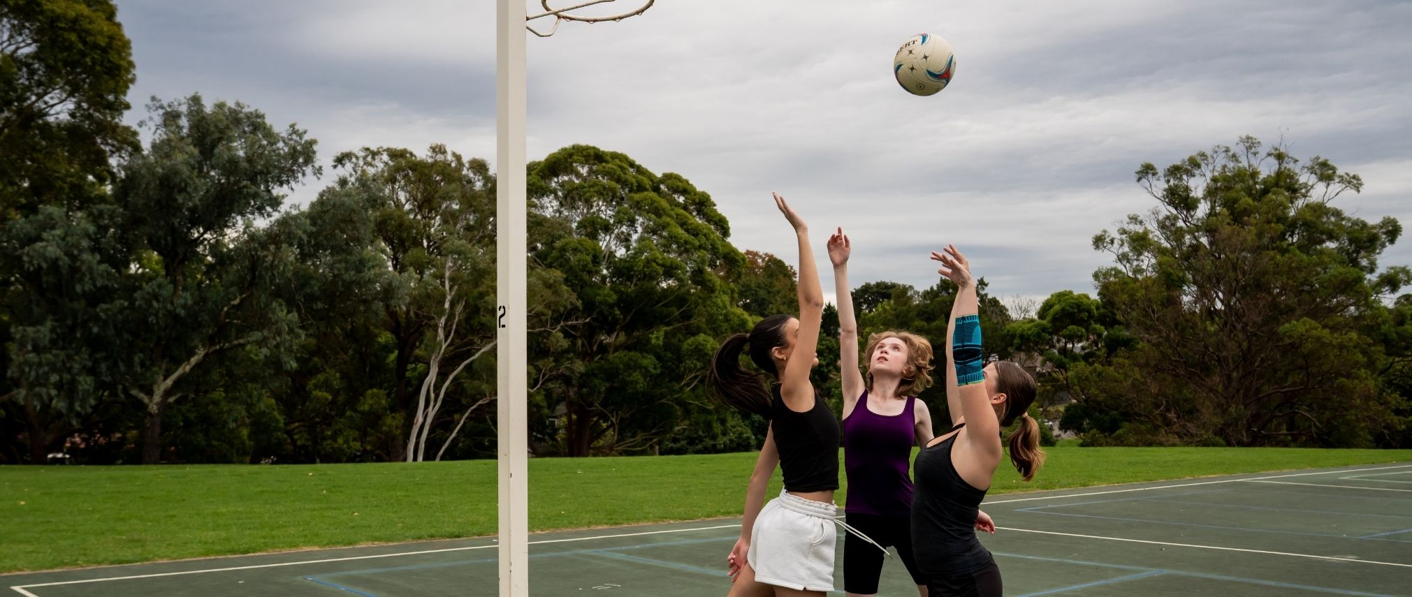 3 girls playing netball. One is shooting the ball into the hoop an 2 are trying to block it. The one throwing the ball is wearing Bauerfeind's Sports Elbow Support