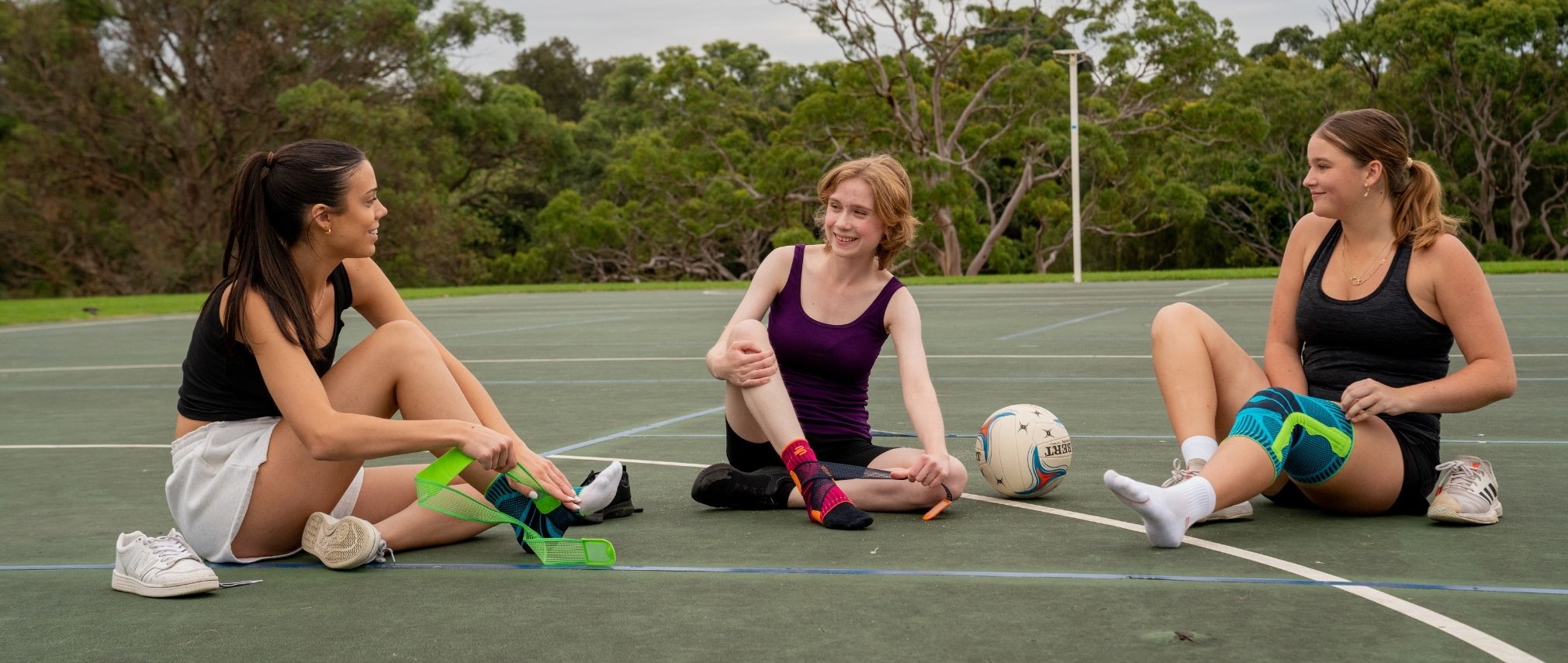 3 girls sitting on a netball court with a netball ball between them. One if them is pulling on a knee brace an the other 2 are strapping on Bauerfeind's Sports Ankle Supports.
