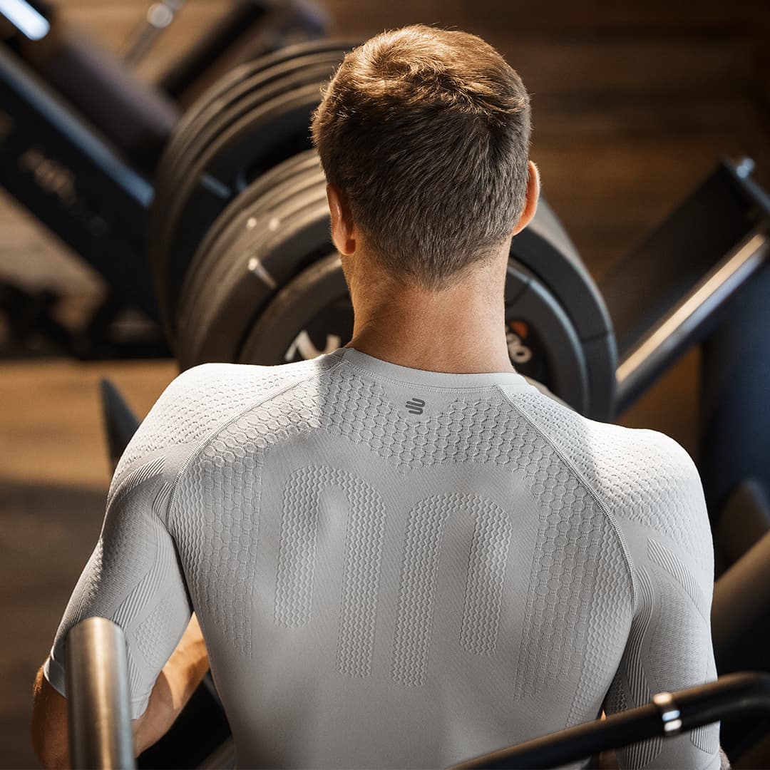 Man from behind using a treadmill in a gym setting wearing a Bauerfeind compression shirt.