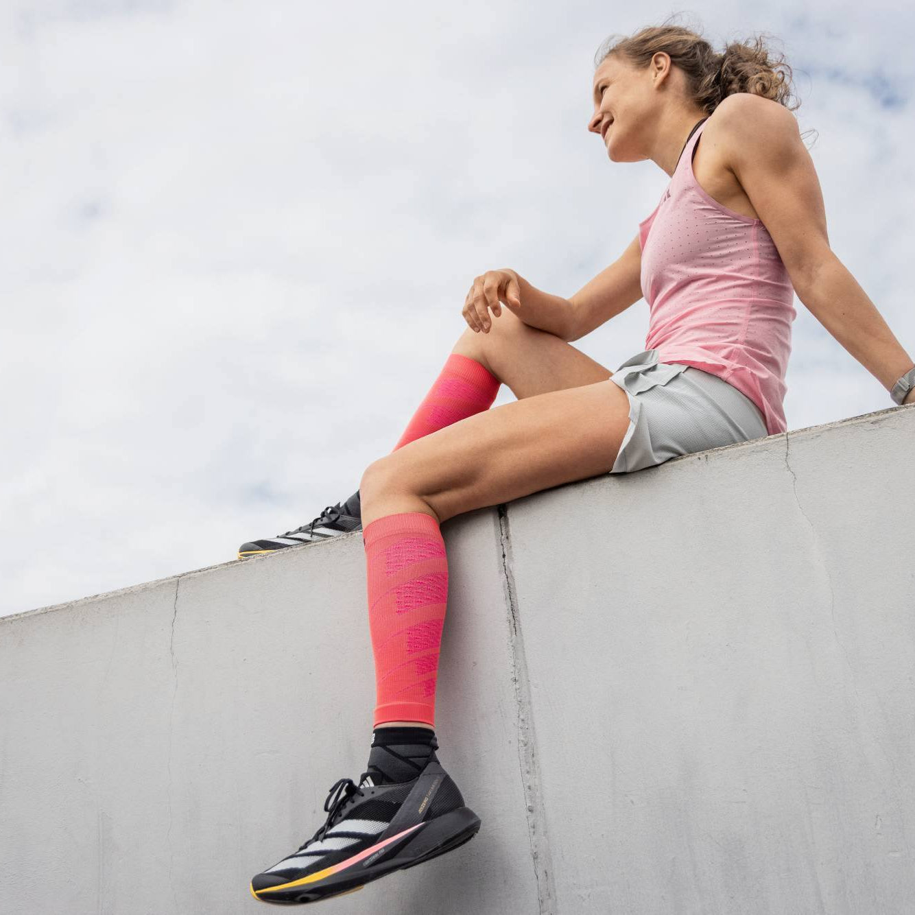 Woman sitting on a ledge wearing pink leggings and black sneakers with a cloudy sky background