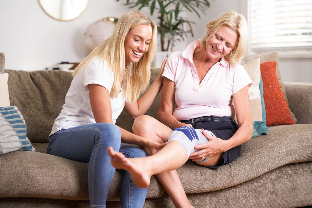 Two women sitting on a couch, one slipping on a Bauerfeind GenuTrain A3 knee support.