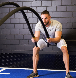 Man exercising with battle ropes against a brick wall, wearing white Bauerfeind Sports Compression Thigh Sleeves.