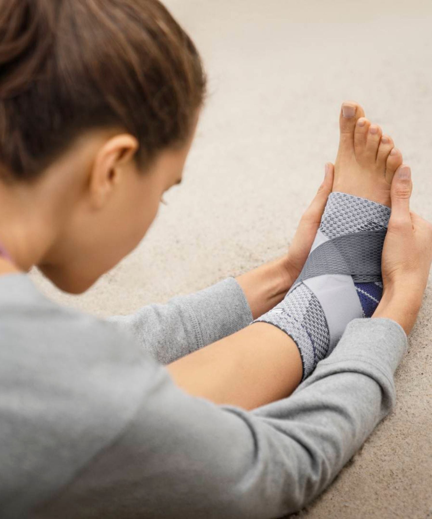 Person stretching with a grey Bauerfeind MalleoTrain Plus ankle support on a carpeted floor.