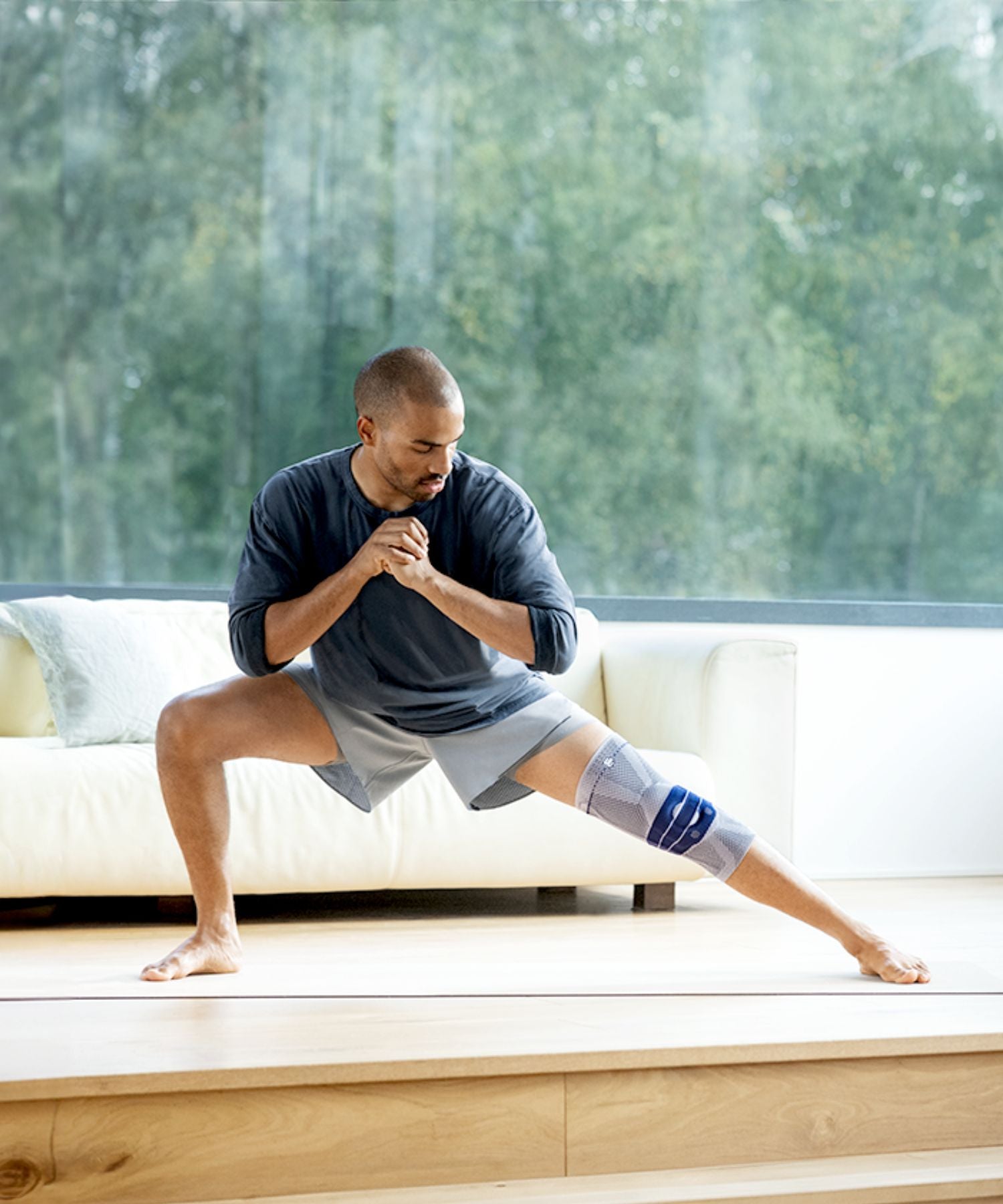 Man stretching with a Bauerfeind GenuTrain knee support on a wooden floor with a blurred green outdoor background.