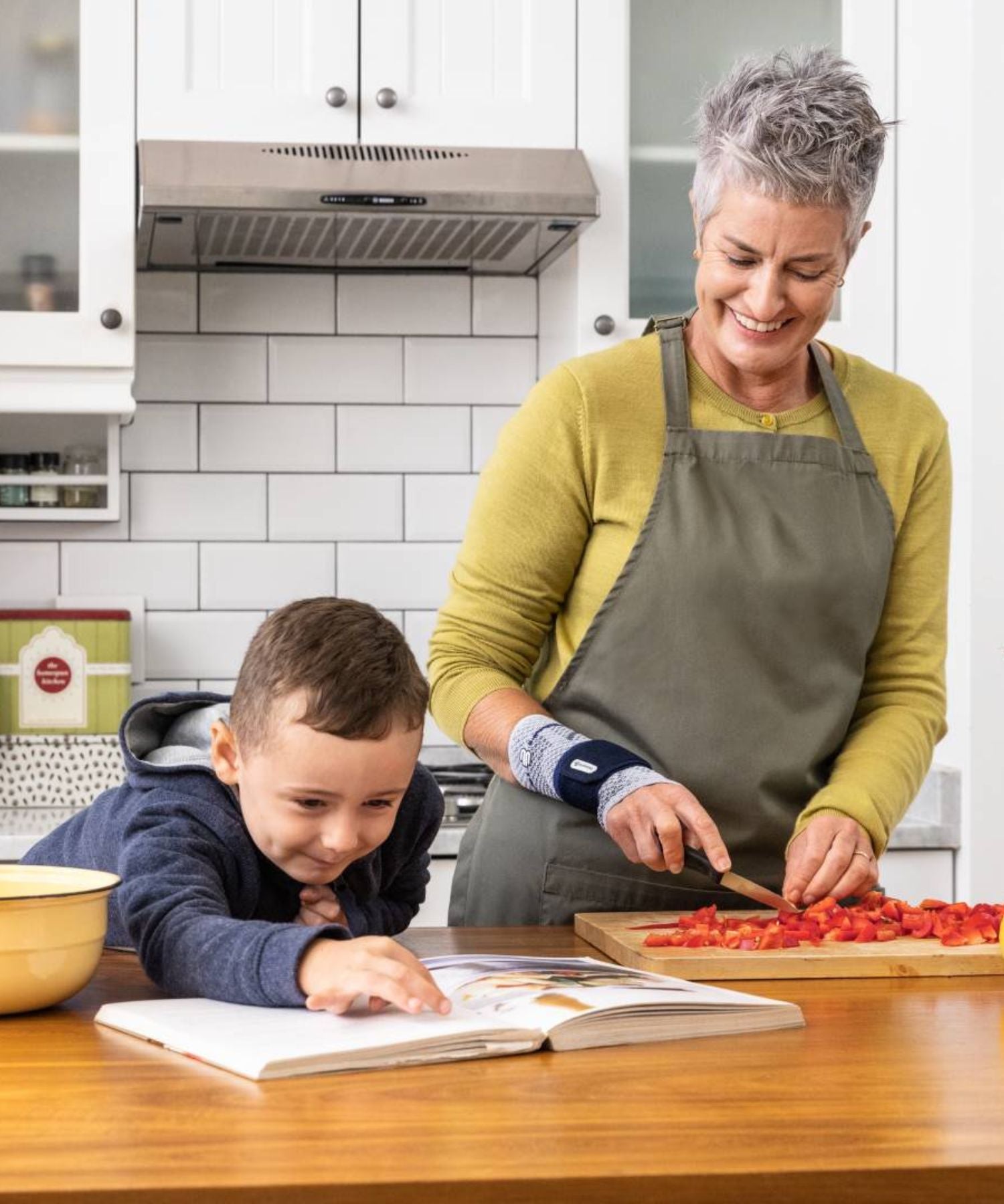 Woman and child in a kitchen, with the woman cutting vegetables and the child looking at the cookbook. She is wearing a Bauerfeind ManuTrain wrist support.