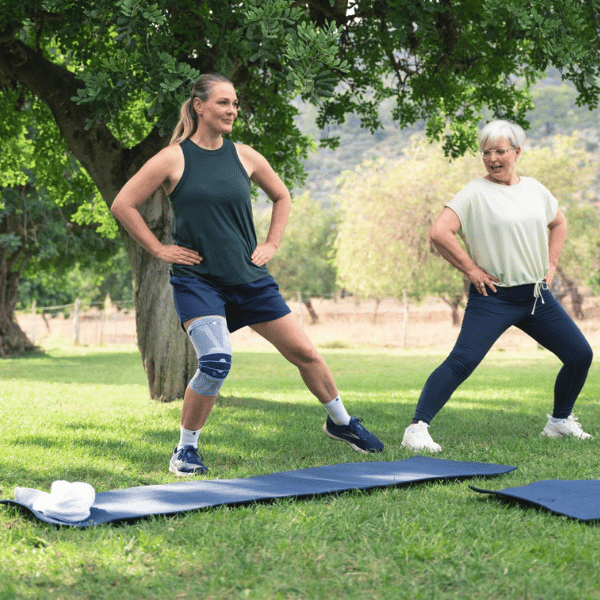 Two women exercising outdoors on grass with trees in the background, on wearing a Bauerfeind GenuTrain A3 knee support.