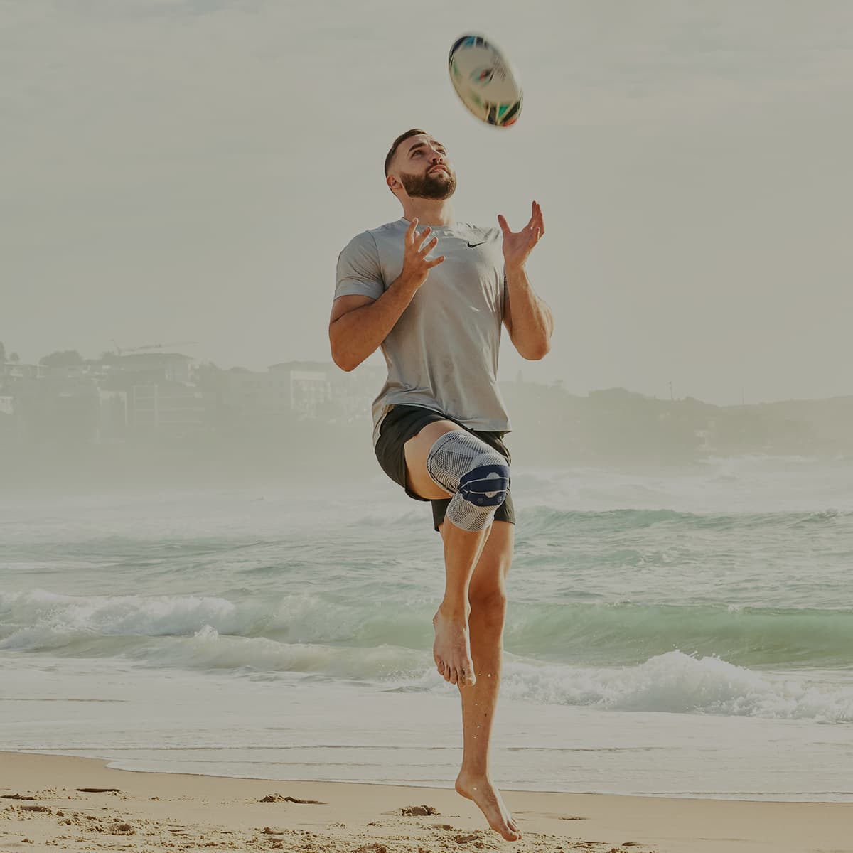 A man on a beach throwing a rugby ball wearing a Bauerfeind GenuTrain knee support