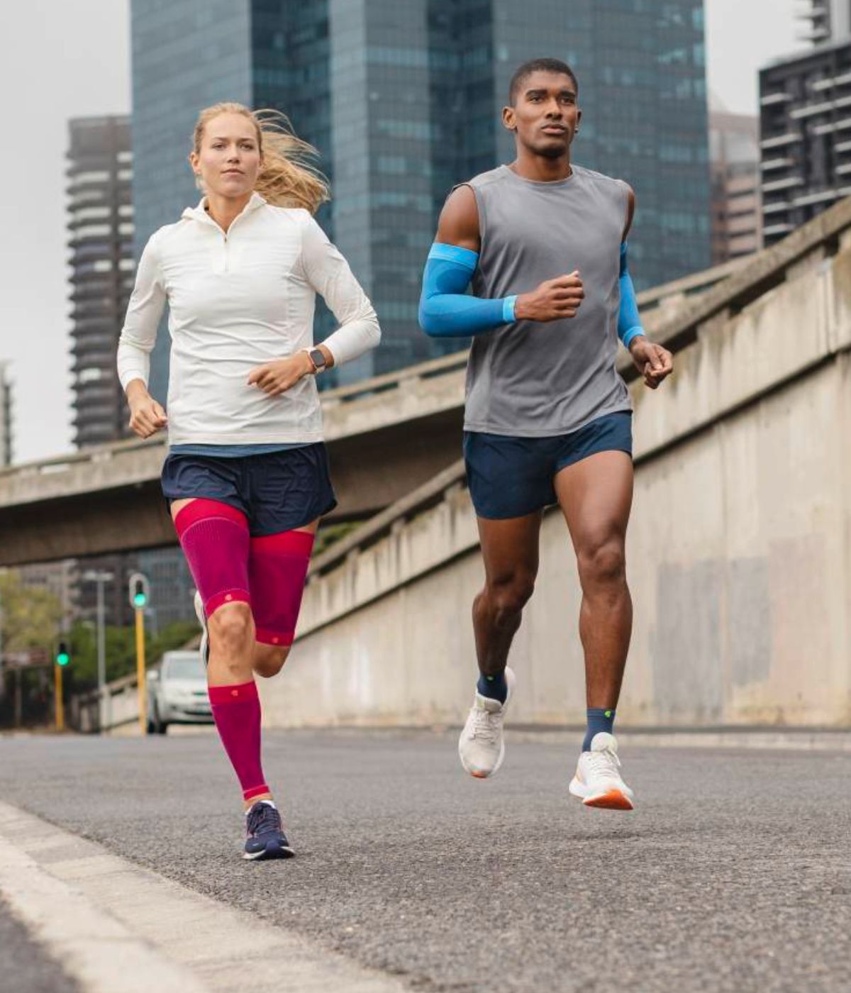 Two runners, a man and a woman, jogging on a city street with tall buildings in the background. Both are wearing colourful Bauerfeind sports compression sleeves.