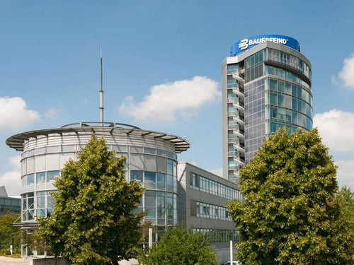 Bauerfeind HQ in Germany. It is 2 buildings, including a small skyscraper with the BAUERFEIND logo at the very top and two shorter buildings that are the production facilities. In front of them are green trees and behind blue skies.