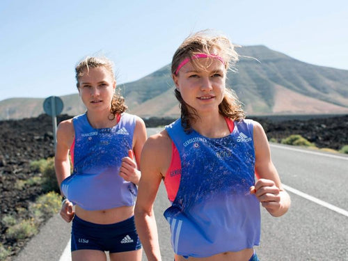 Hahner twins running on a road with mountains in the background. The twins are blonde women wearing bright orange sports bras, blue jerseys, and dark blue shorts.