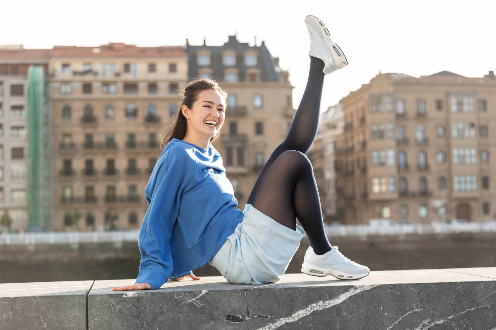 Woman sitting on a ledge with one leg raised in an urban setting, wearing a Bauerfeind VenoTrain compression stocking.